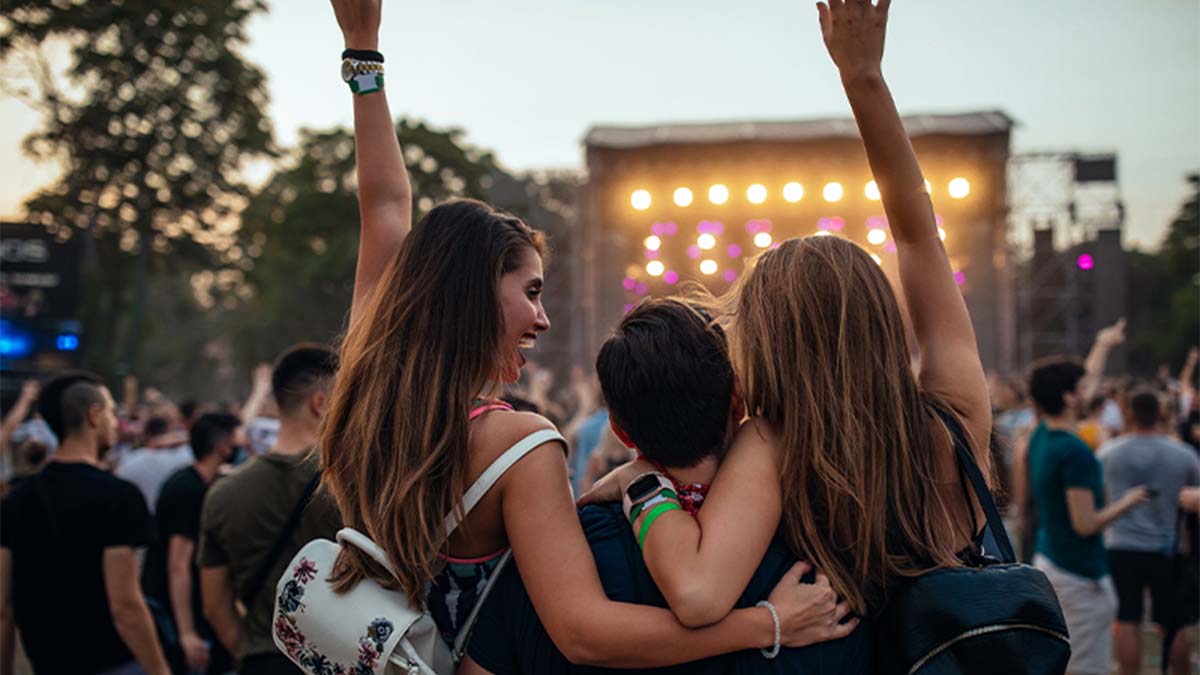 Multidão num concerto ao ar livre; várias pessoas de costas com braços no ar, palco iluminado e público ao fundo.